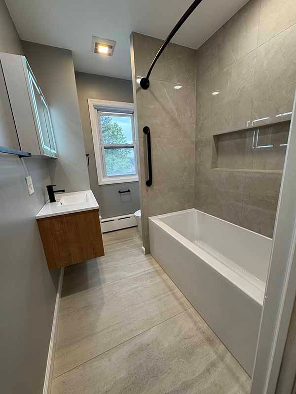 Modern bathroom with gray tile, wooden vanity, and a tub with a safety grab bar.