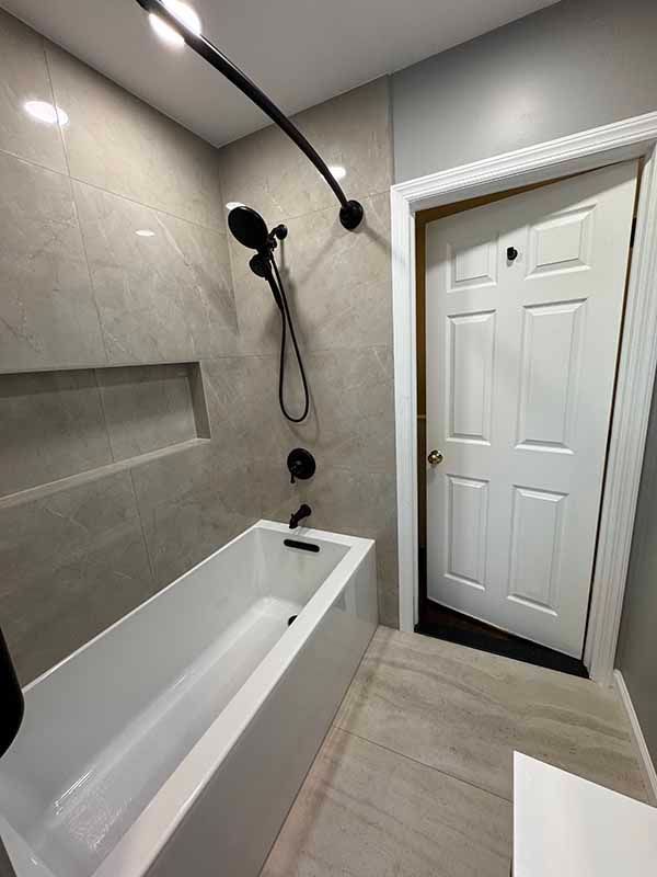 Modern bathroom with a white bathtub, black showerhead, and a white door. Gray tiles on the walls and floor.