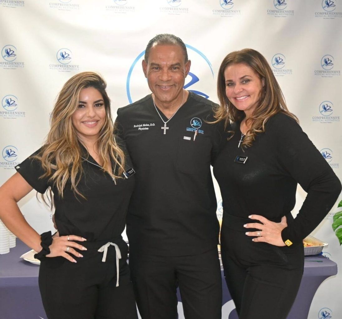 Three people smiling, wearing black scrubs, posing in front of a blue logo background.