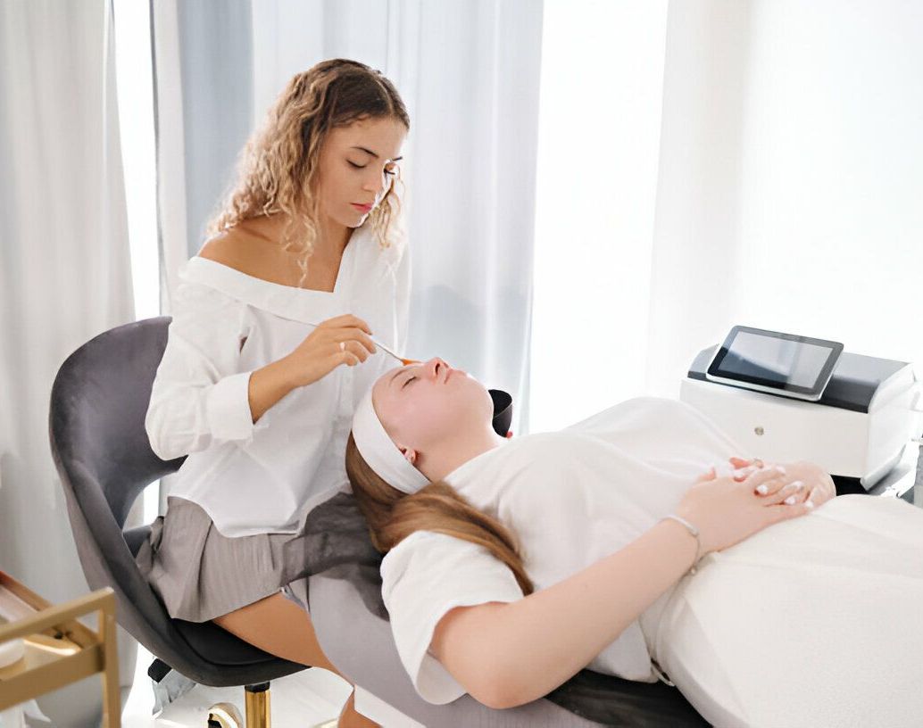 Woman giving facial treatment to another woman lying down. Bright room with medical equipment.