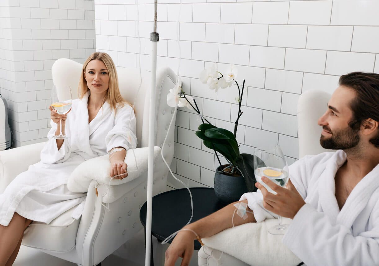 Woman and man in white robes receiving IV drips in a bright room.
