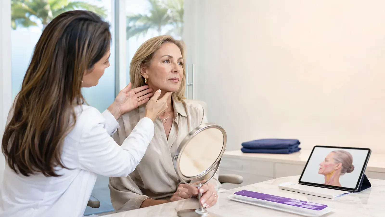 Woman receives a facial consultation while looking in a hand mirror beside a tablet displaying a face diagram
