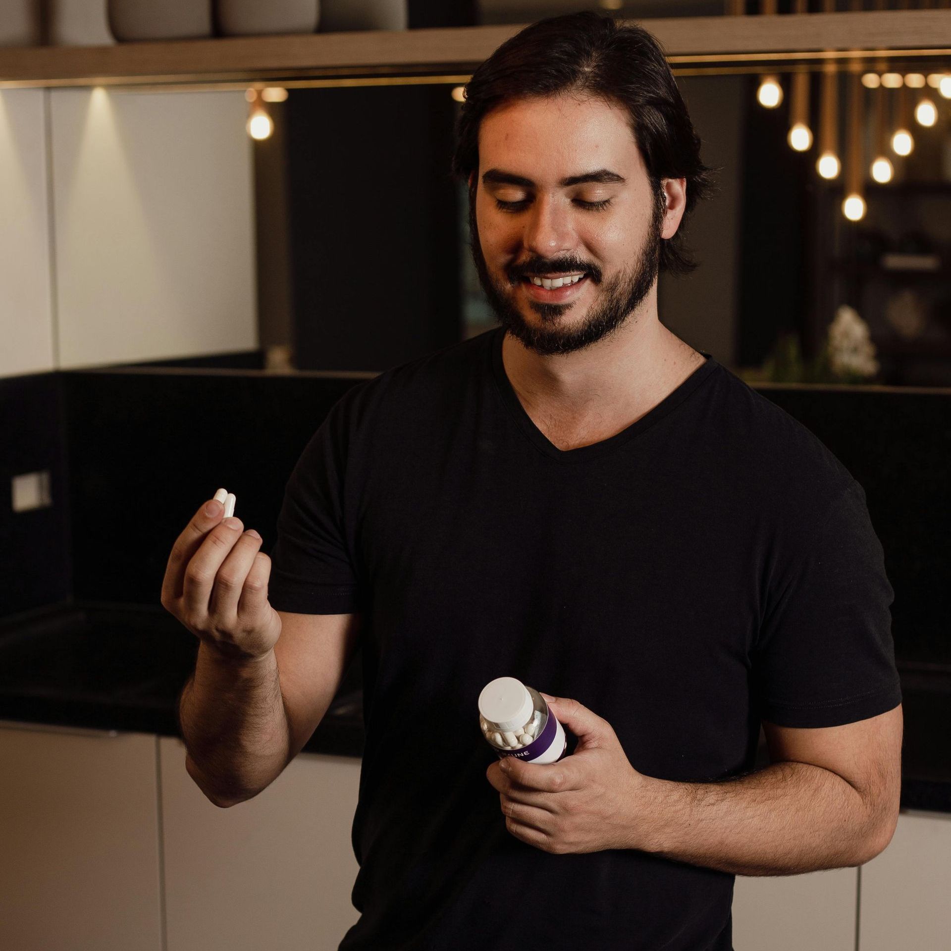 Man holding a pill from a bottle, smiling. Black shirt, modern kitchen background.