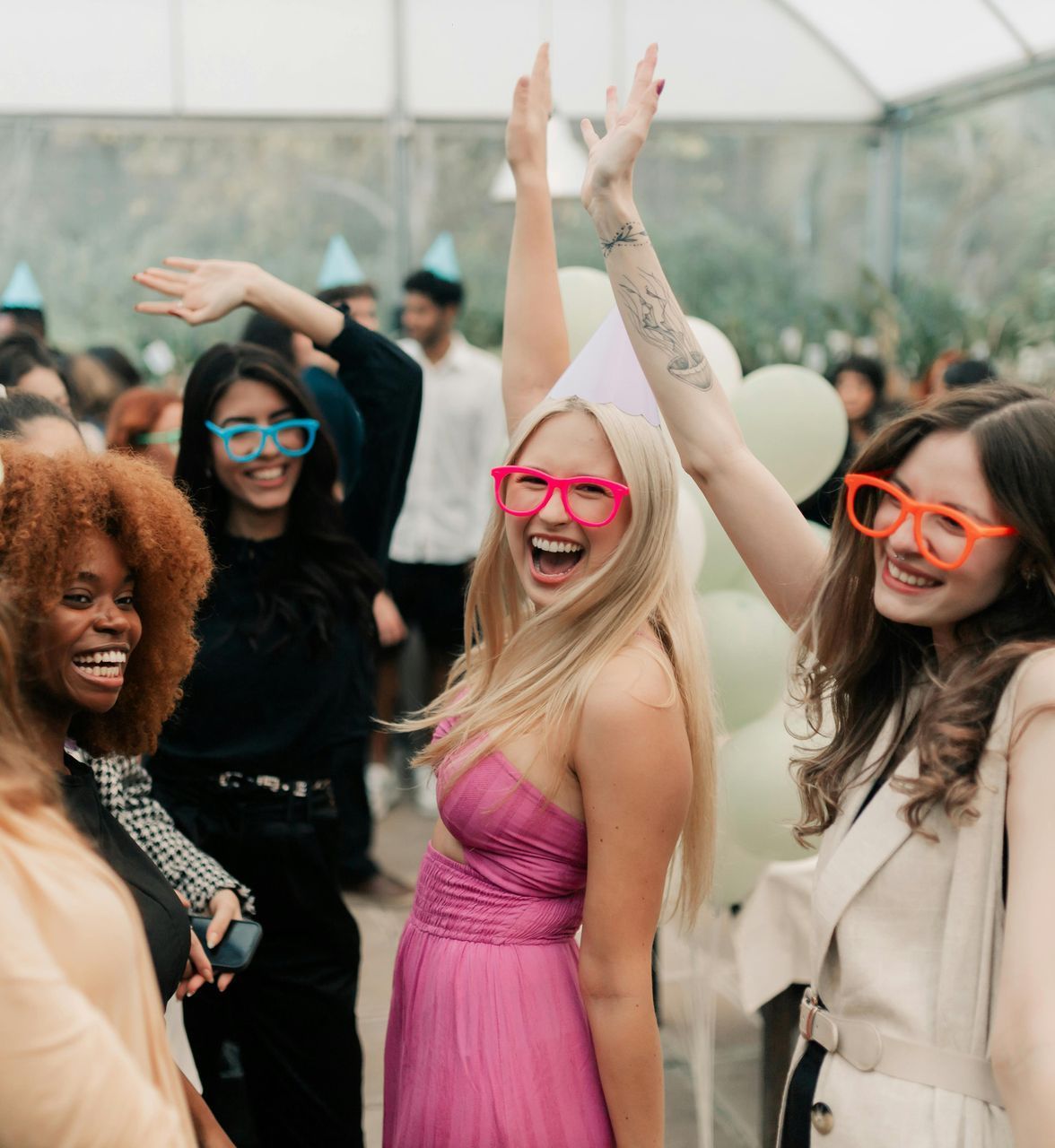 Group of people celebrating, wearing party hats and novelty glasses, raising hands and smiling.