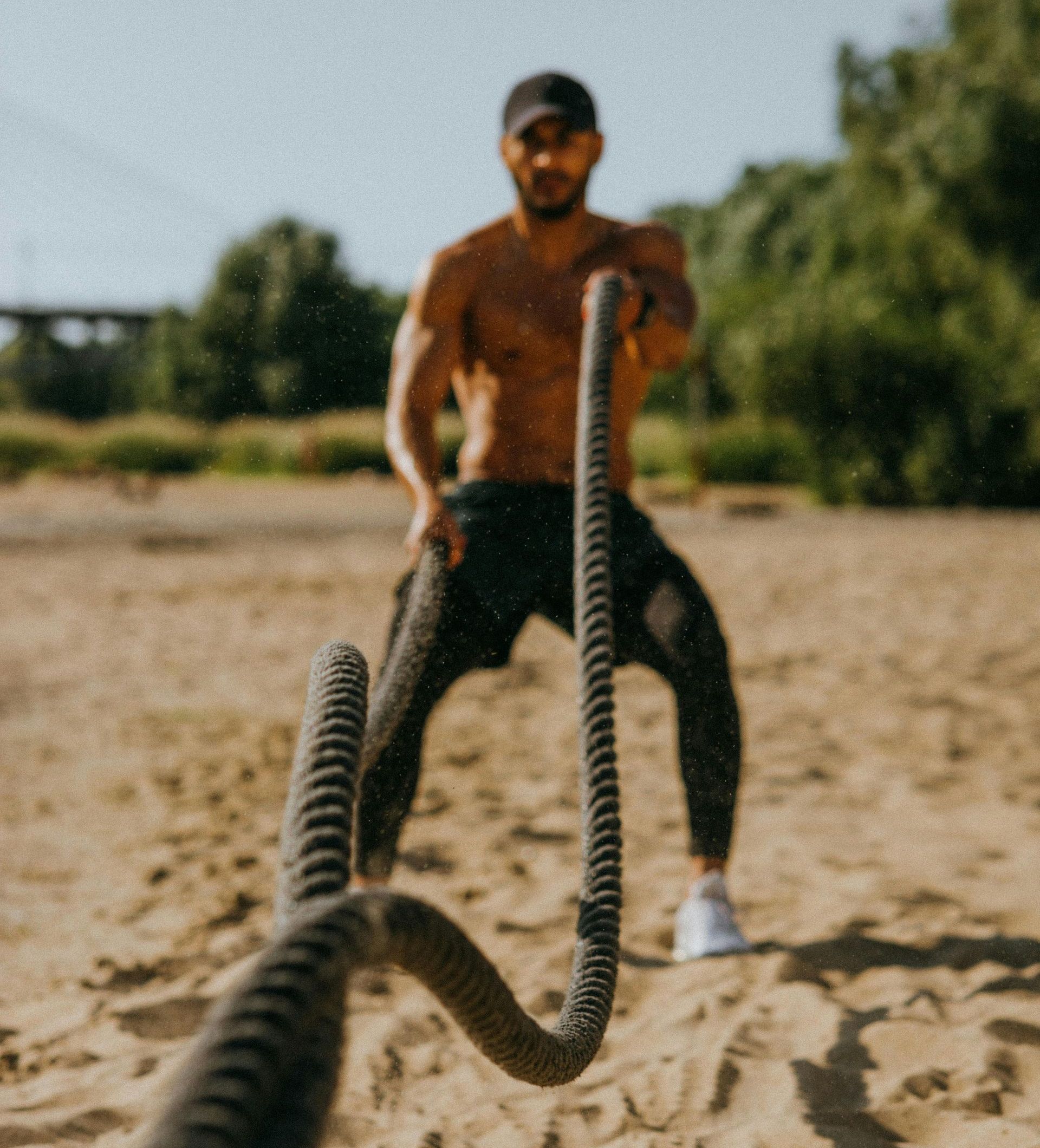 Man doing battle rope exercises outdoors on a sandy surface.