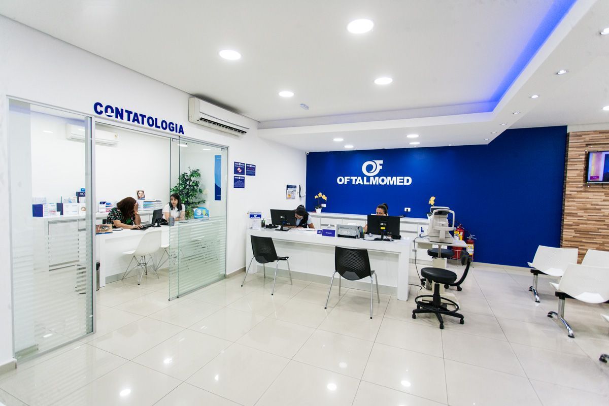 Recepção de consultório médico bem iluminada, com funcionários em mesas brancas, parede azul com a placa