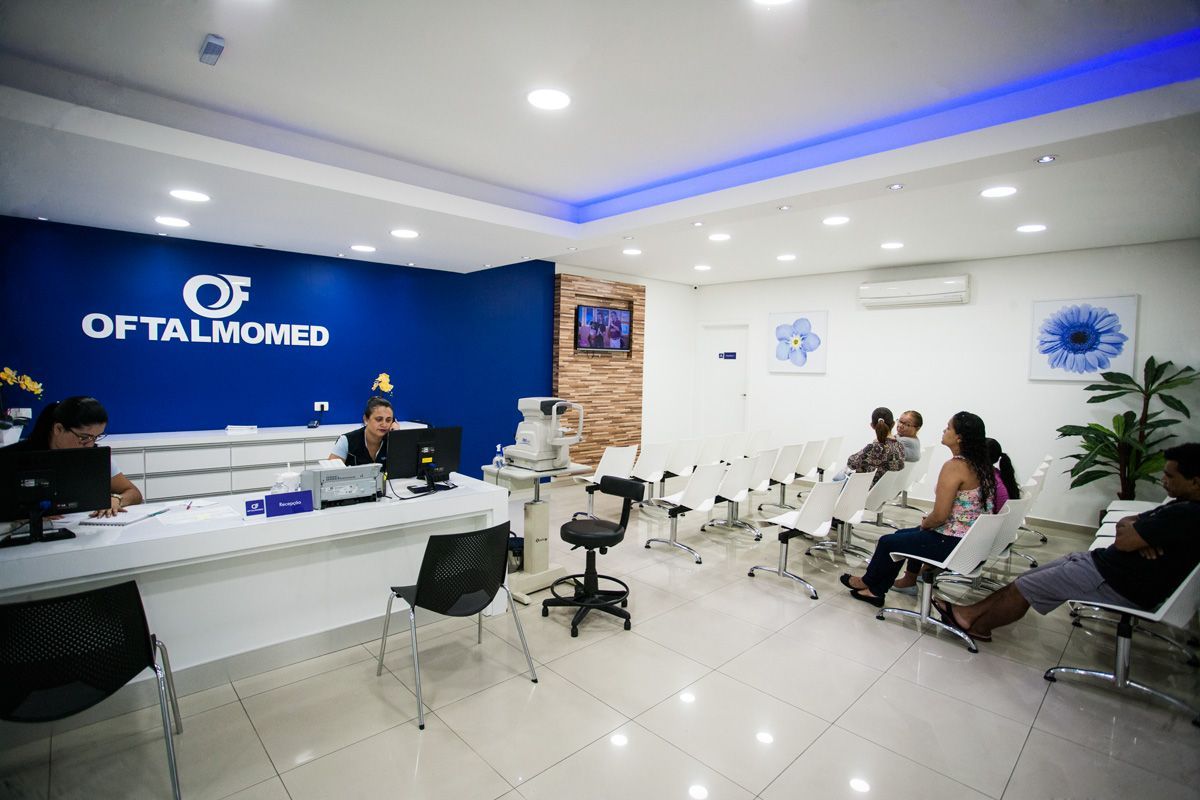 Área de recepção em uma clínica oftalmológica com funcionários atrás de um balcão e várias pessoas aguardando sentadas em cadeiras.