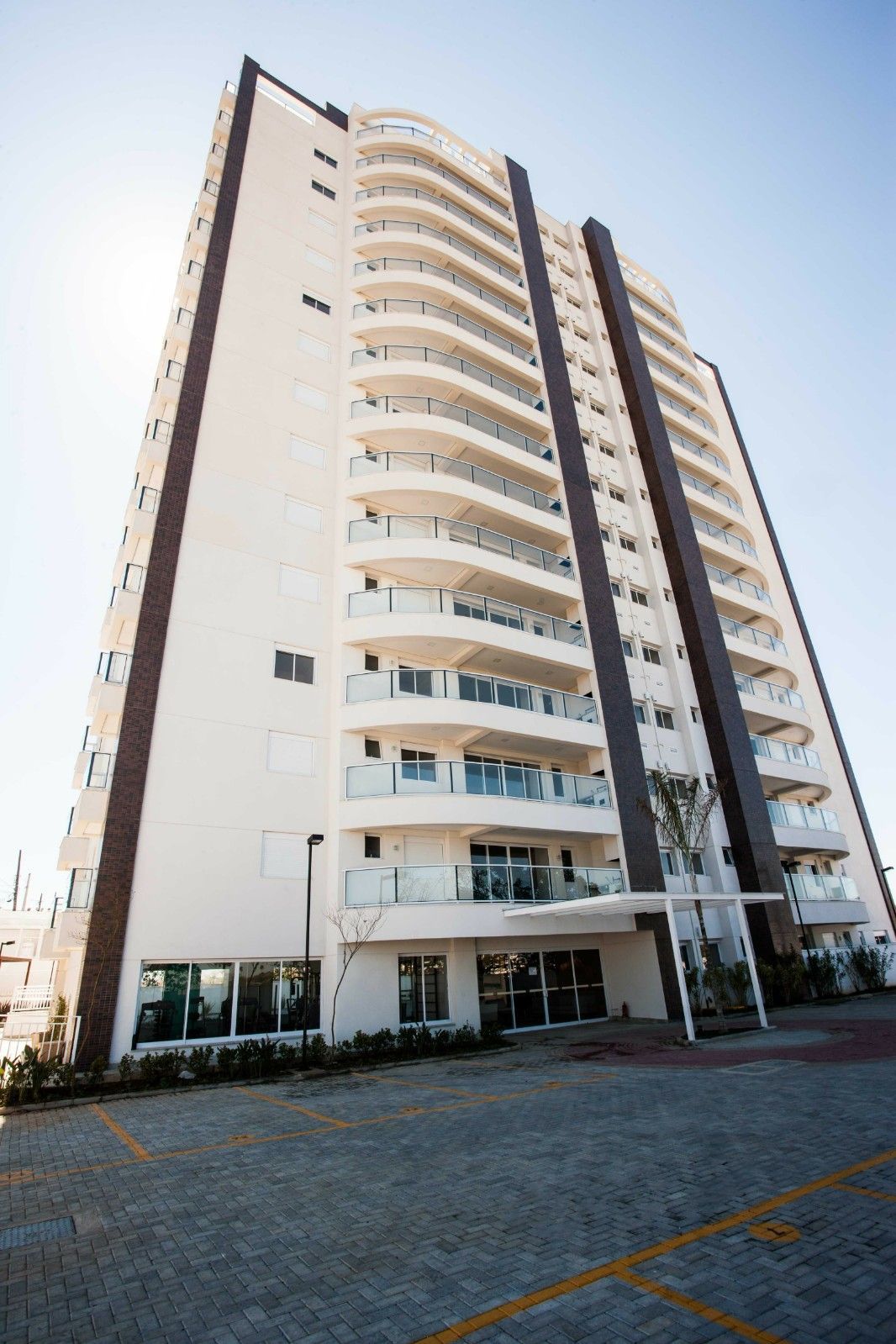 Um prédio de apartamentos moderno, de cor clara, com varandas curvas, detalhes escuros e uma área de entrada pavimentada.