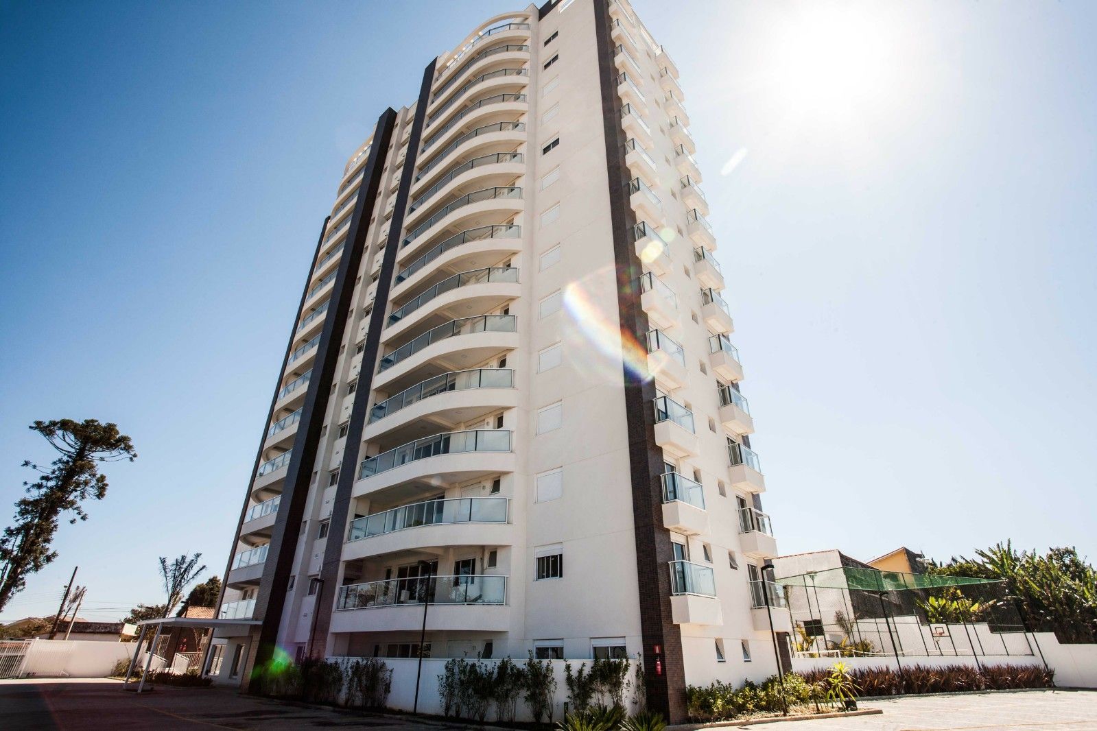 Um alto e moderno prédio de apartamentos branco, com varandas curvas de vidro, destaca-se contra um céu azul brilhante em um dia ensolarado.