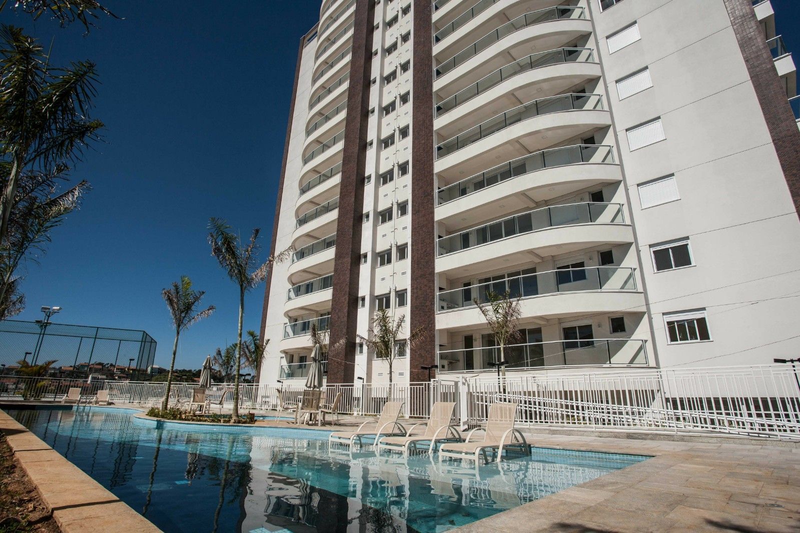 Um prédio residencial de vários andares com varandas ao lado de uma piscina externa em um dia claro e ensolarado.