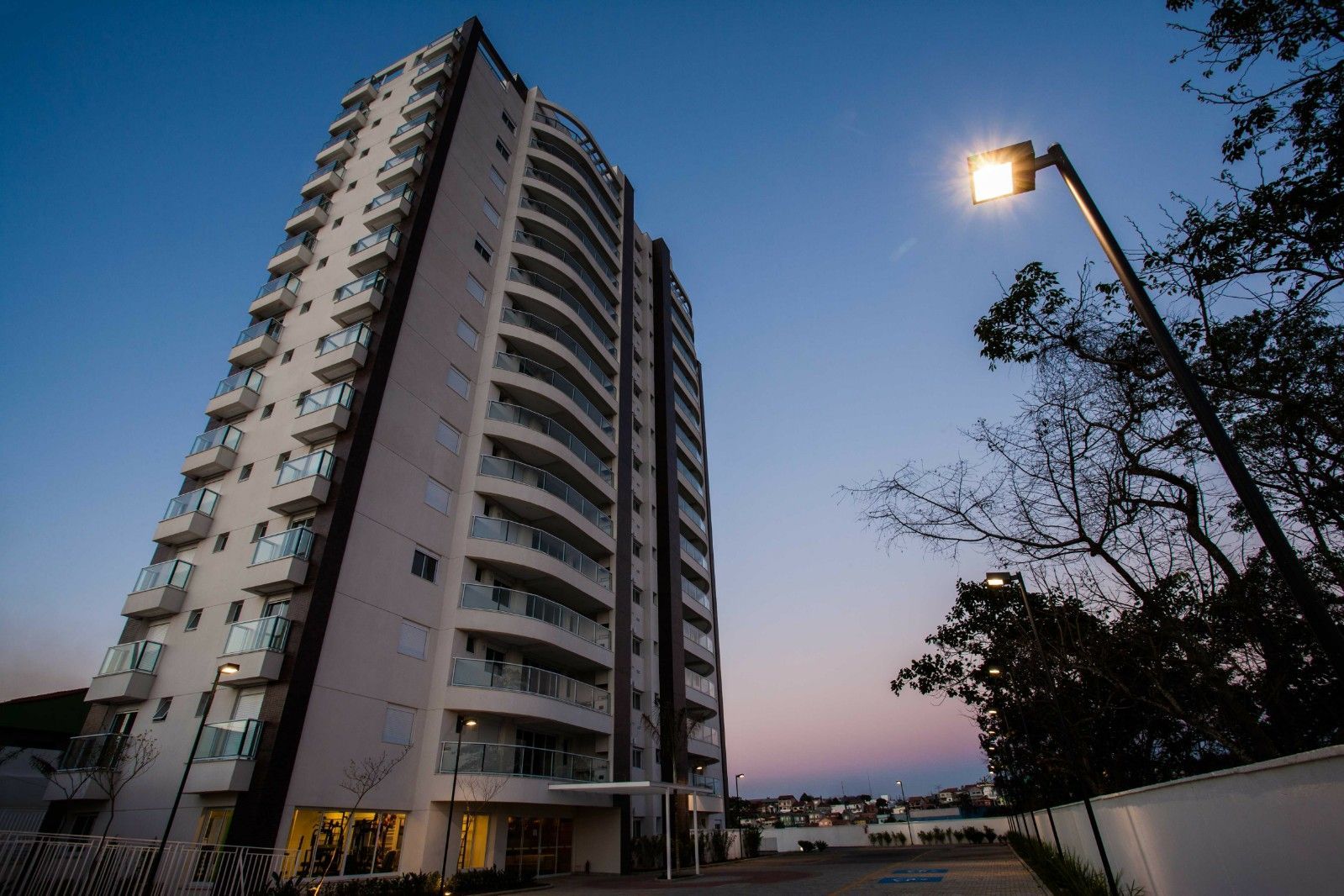Um moderno prédio de apartamentos de vários andares com varandas se destaca contra um céu crepuscular, iluminado por um poste de luz próximo.