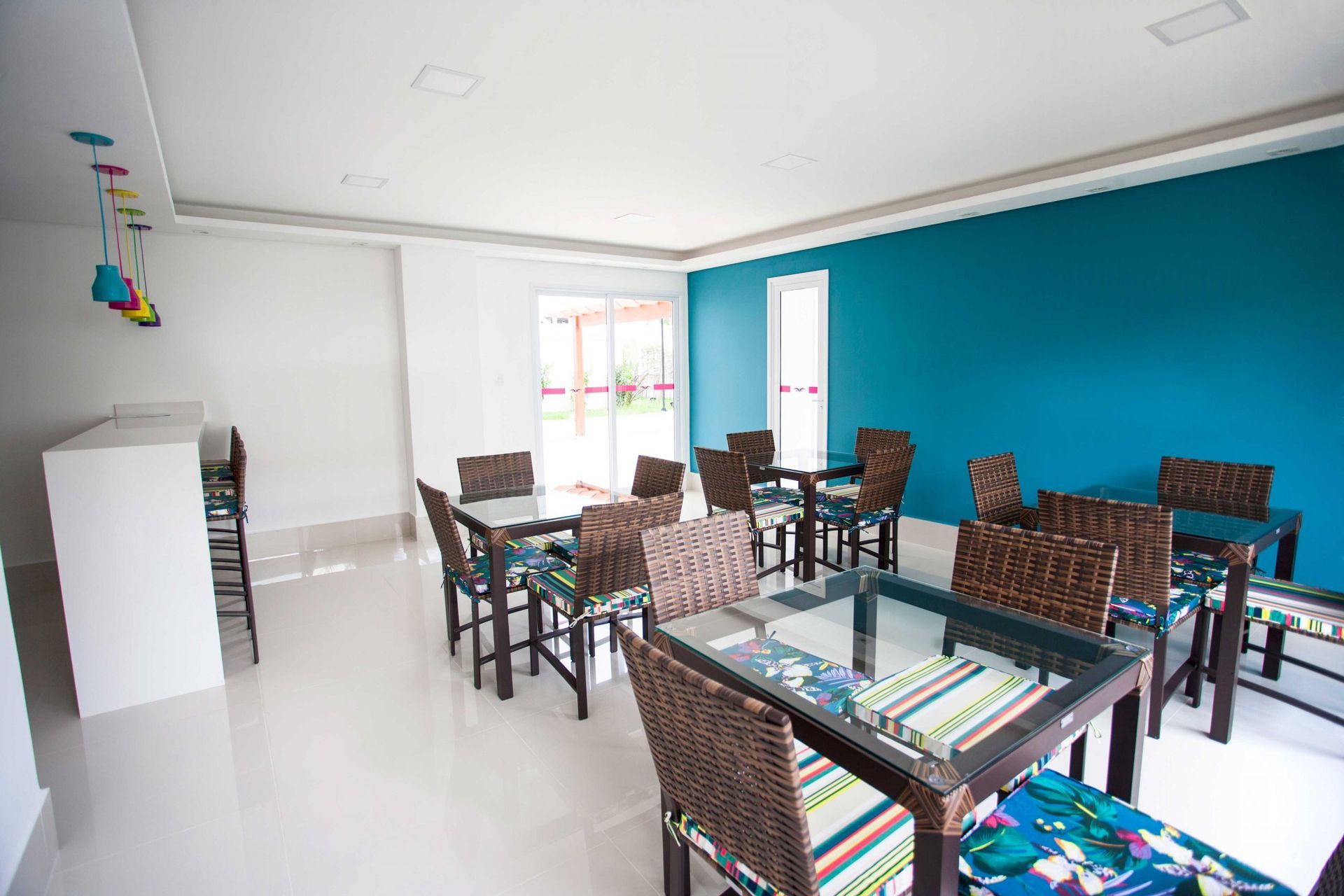 Uma sala de jantar iluminada com mesas de vidro, cadeiras de vime, um balcão de serviço branco e uma parede de destaque em um tom vibrante de azul.