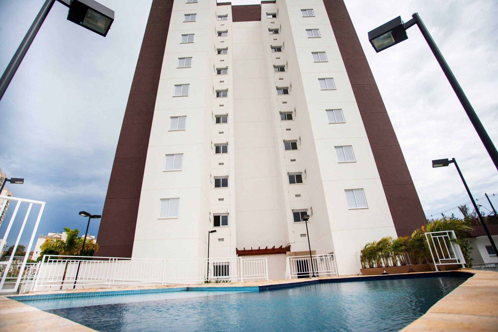 Um prédio de apartamentos alto e moderno, com paredes brancas e marrons, ergue-se acima de uma piscina no quintal, sob um céu nublado.
