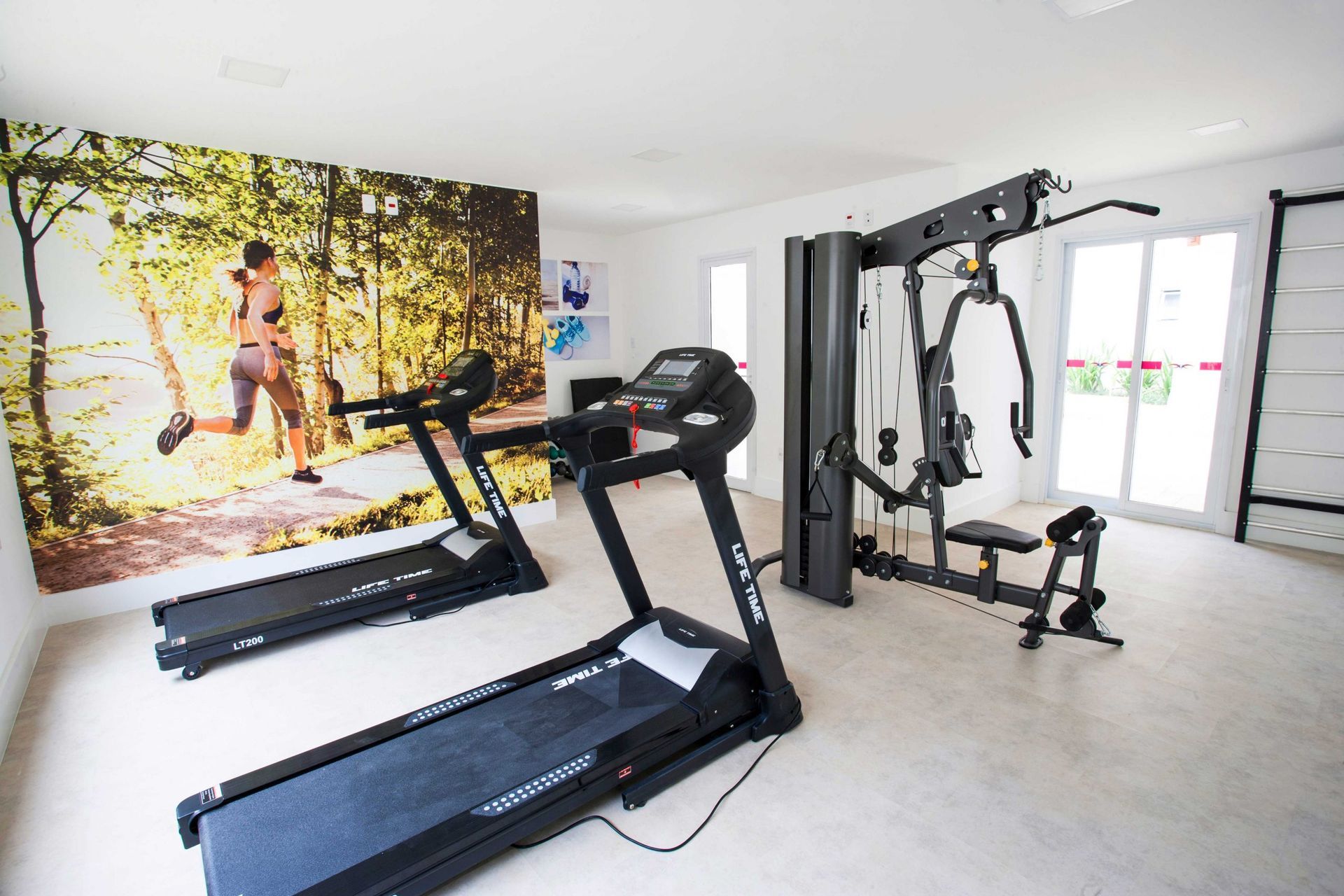 Uma academia residencial bem iluminada com duas esteiras em frente a um mural na parede com a imagem de uma pessoa correndo e um aparelho de musculação multifuncional nas proximidades.