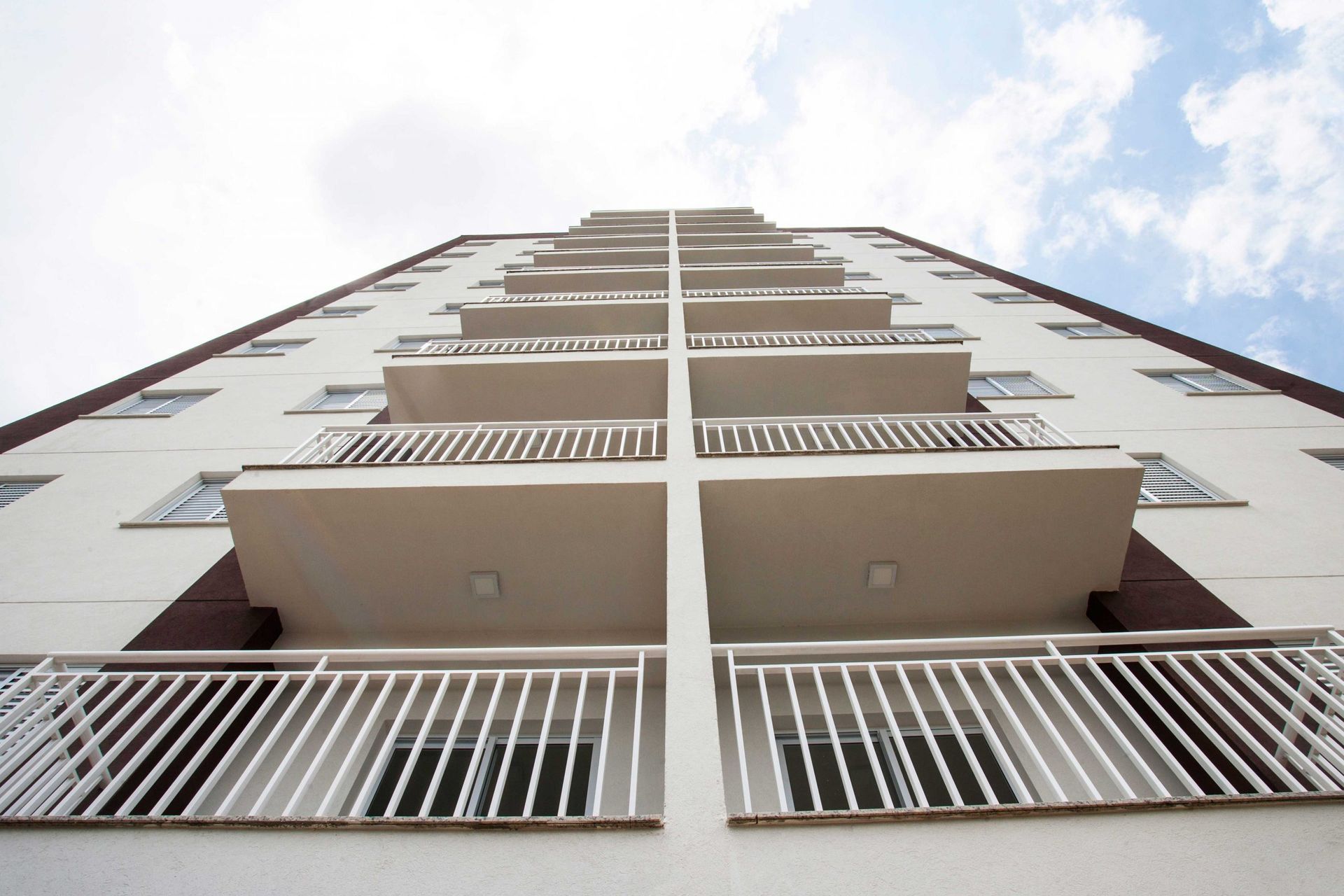 Vista em ângulo baixo da fachada de um moderno prédio de apartamentos branco com varandas simétricas contra um céu azul e nublado.