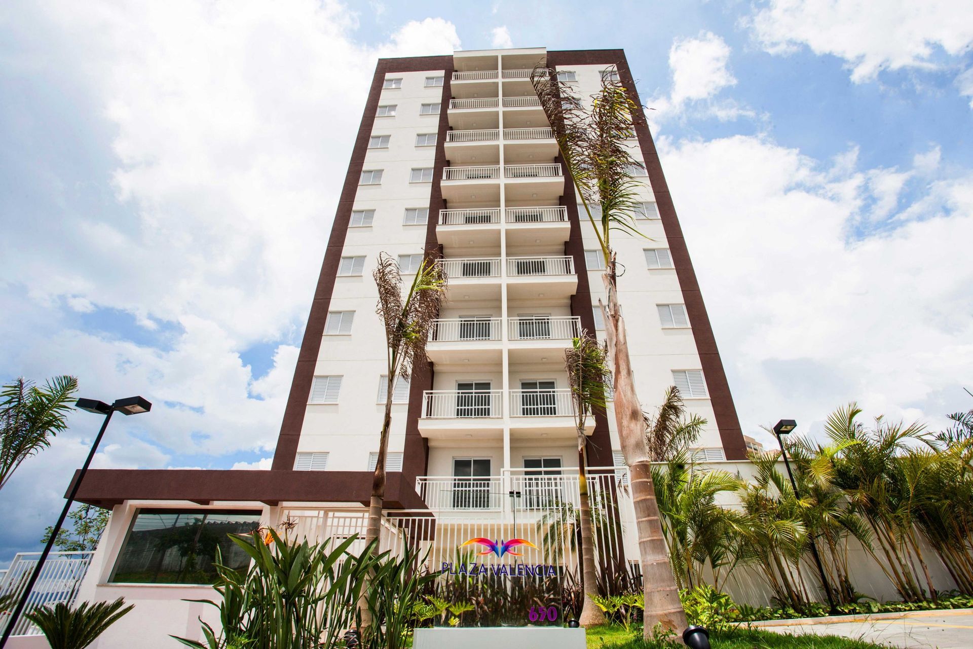 Um moderno edifício residencial de vários andares com varandas brancas e detalhes em marrom, emoldurado por palmeiras contra um céu nublado.