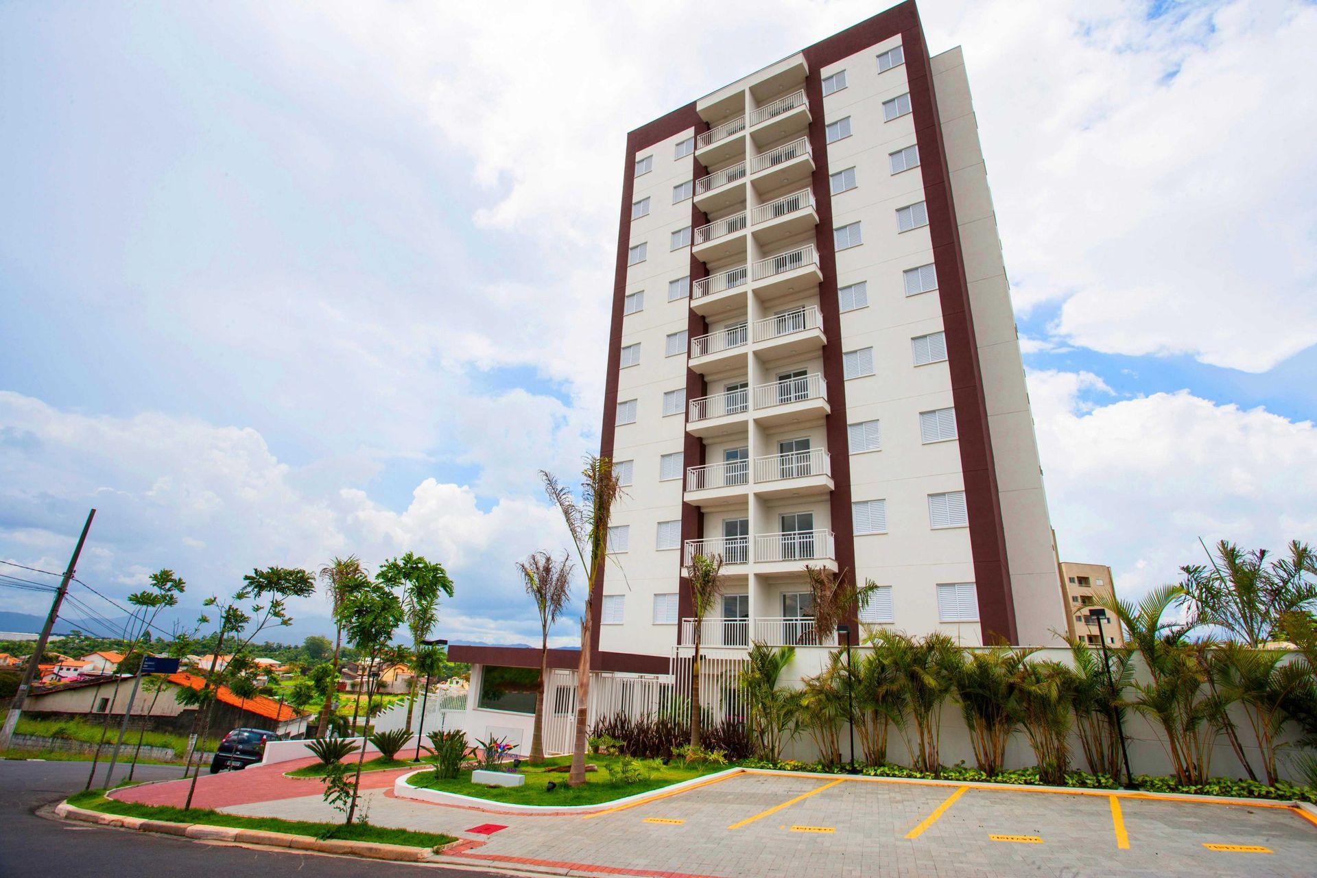 Um prédio de apartamentos moderno, branco, com vários andares, painéis laterais marrons, varandas e um estacionamento adjacente.