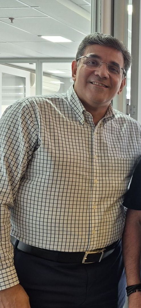 Uma pessoa sorridente usando óculos e uma camisa xadrez clara de botões, por dentro de calças escuras.