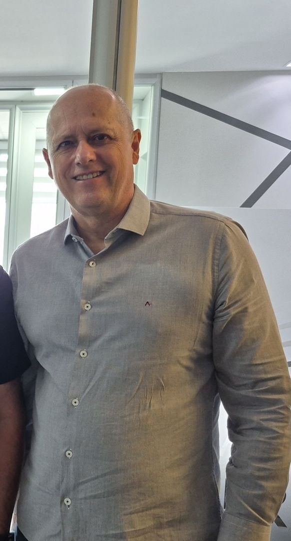 Uma pessoa sorridente, vestindo uma camisa cinza-clara de botões, está em pé em um escritório bem iluminado.