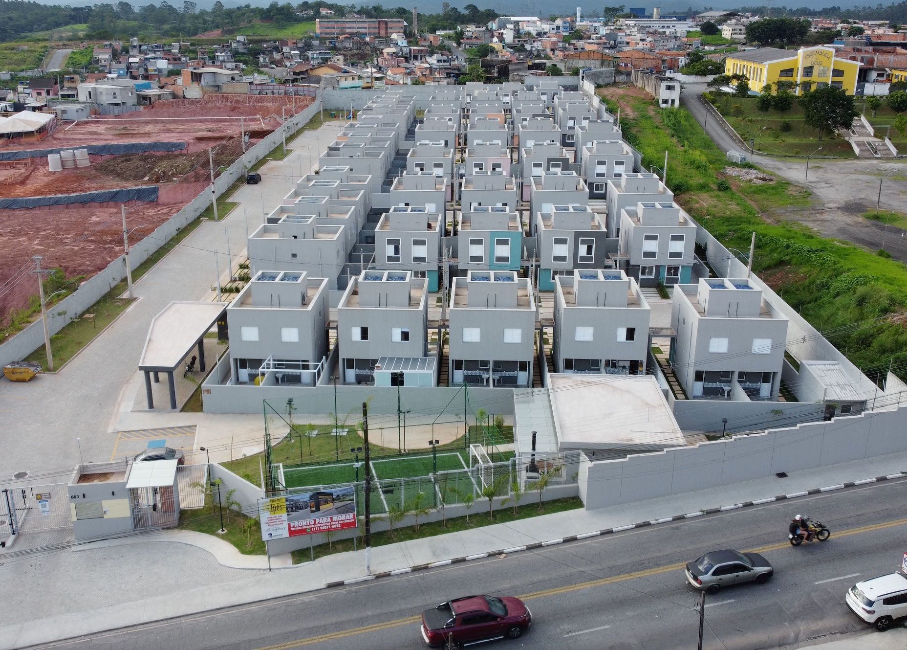 Vista aérea de um moderno complexo residencial de casas geminadas, com fileiras de residências cinzentas, um pequeno campo de futebol e uma entrada com portão.