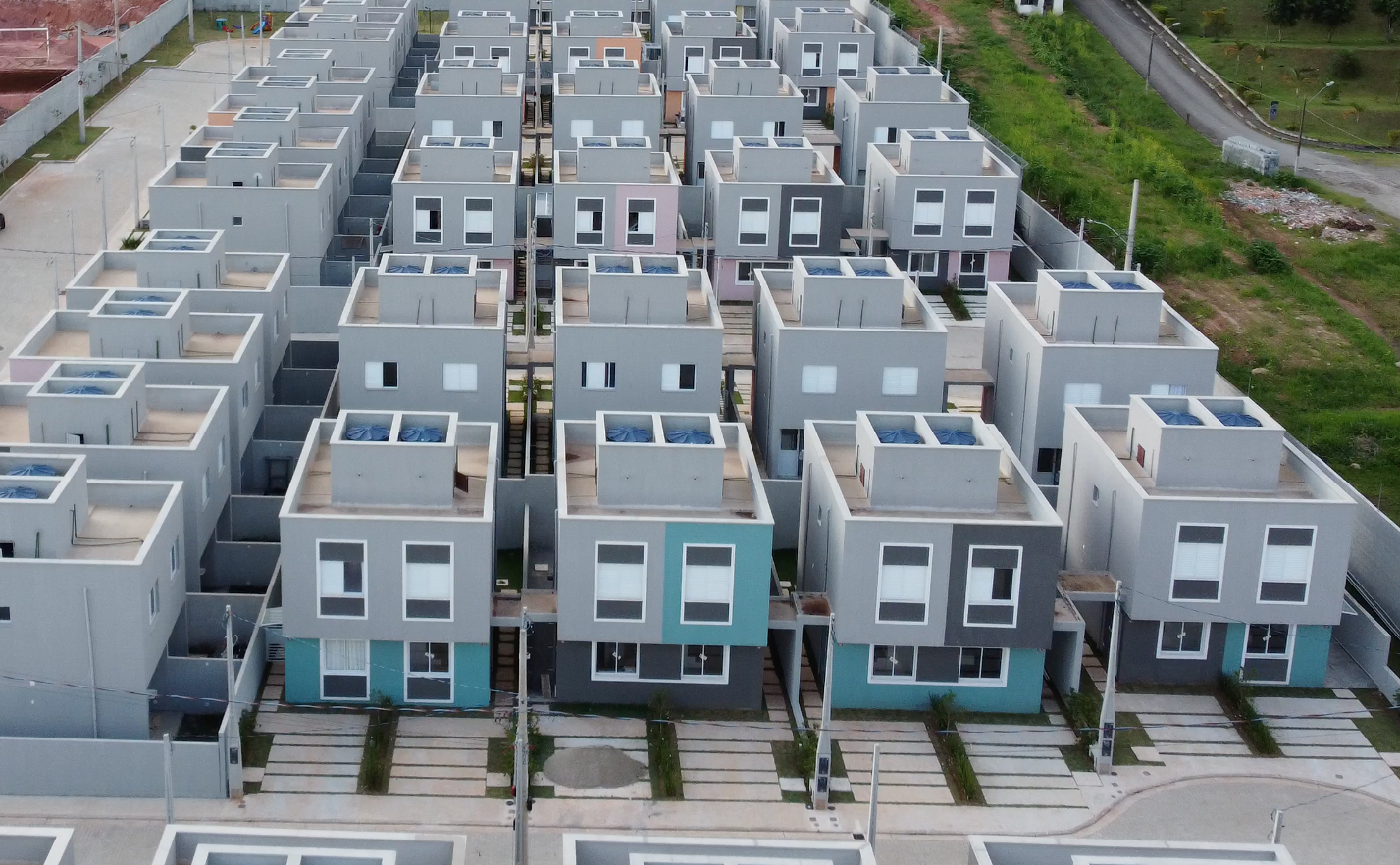 Vista aérea de um denso e moderno conjunto habitacional suburbano com fileiras de casas cúbicas idênticas de dois andares, em tons de cinza e azul.