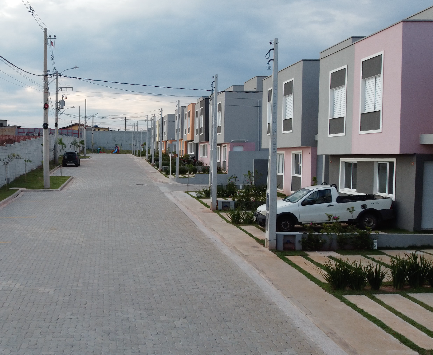 Uma fileira de casas geminadas modernas de dois andares em vários tons pastel alinha-se numa rua pavimentada sob um céu nublado.