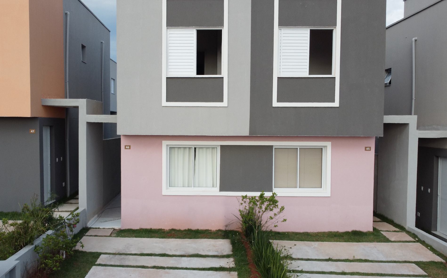 Uma casa de dois andares com o térreo rosa, o andar superior cinza e uma entrada de automóveis pavimentada, localizada entre outras casas semelhantes.