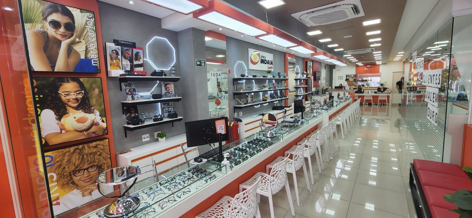 Interior de uma ótica com vitrines, fileiras de óculos e cartazes promocionais com pessoas usando óculos.