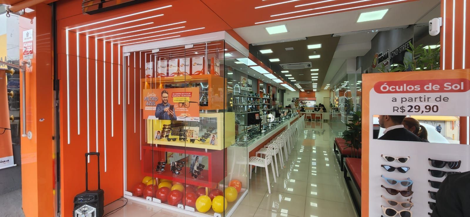 Fachada laranja vibrante de uma ótica, com uma vitrine de óculos de sol e uma placa com os preços.