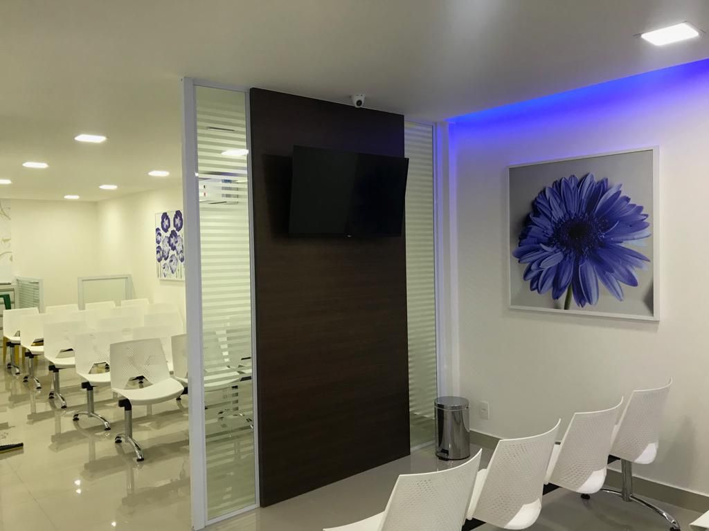 Uma sala de espera limpa com fileiras de cadeiras brancas, uma televisão em uma parede divisória escura e uma grande obra de arte em forma de flor azul.