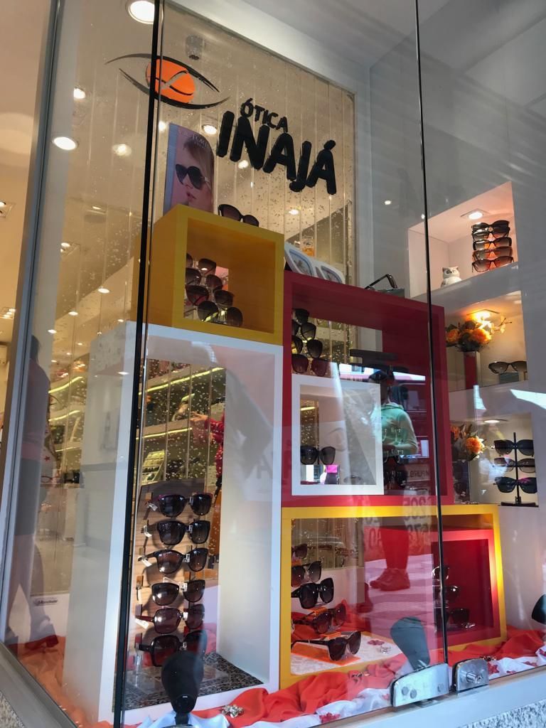 Vitrine da loja Otica Inaja exibindo diversos óculos de sol em prateleiras coloridas nas cores vermelho, amarelo e branco.