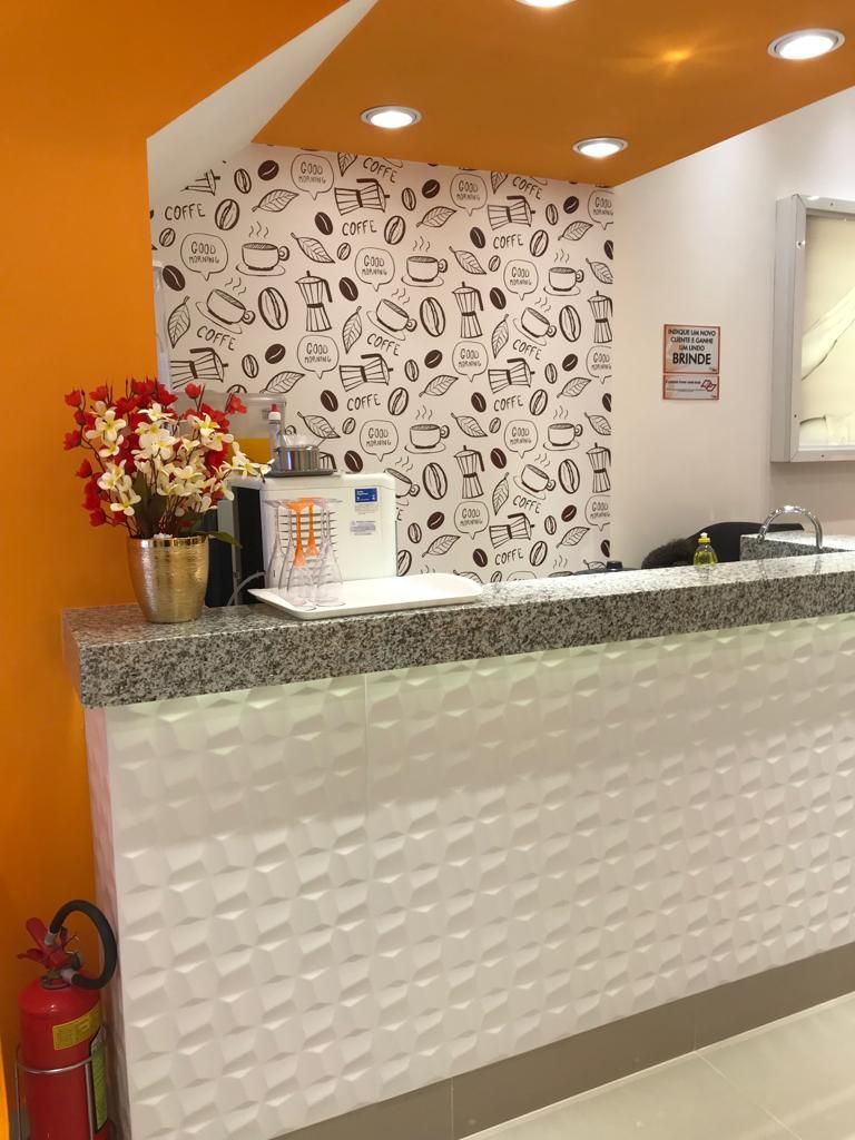 Balcão de recepção com frente branca estampada, papel de parede com tema de café ao fundo, detalhes em laranja no teto e flores.