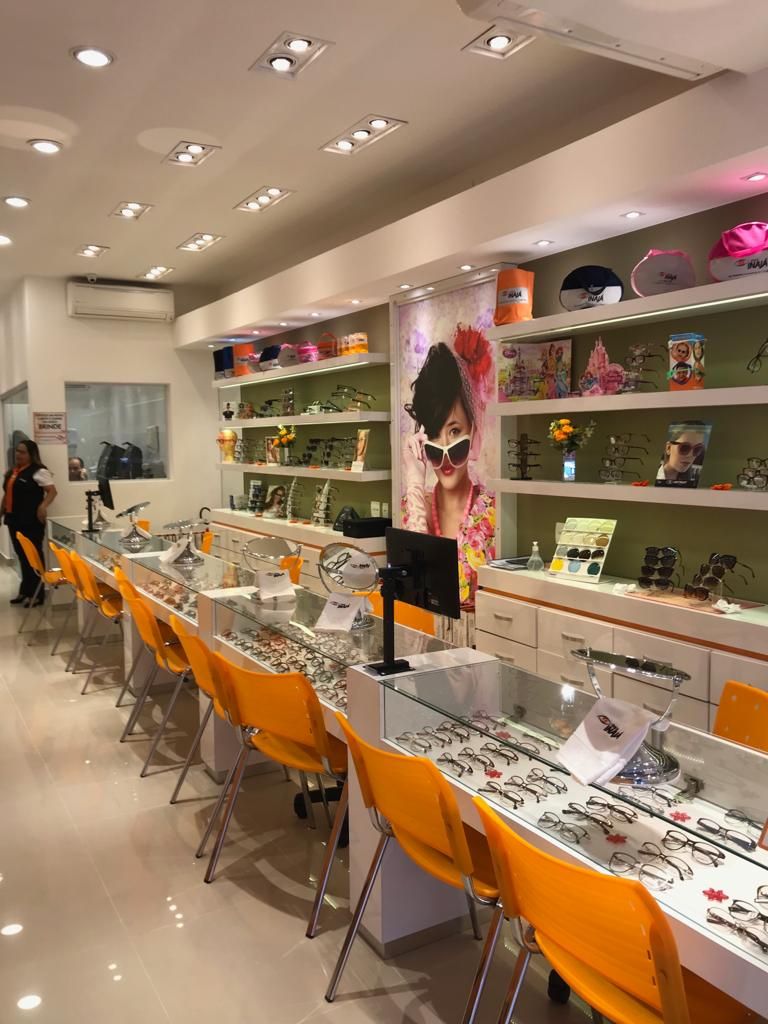 Interior de uma ótica com óculos expostos em prateleiras brancas, balcões de vidro e fileiras de cadeiras laranja.