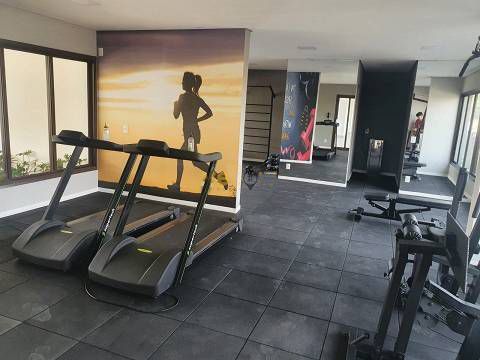 Interior de uma academia com esteiras, um banco de musculação e um grande mural na parede retratando uma pessoa correndo contra um pôr do sol dourado.