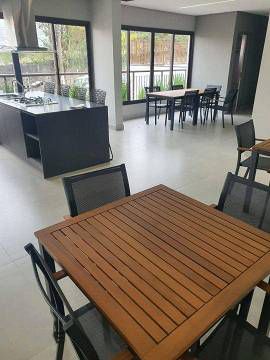 Sala de jantar moderna com uma mesa quadrada de madeira em primeiro plano e uma ilha de cozinha com bancada escura ao fundo.