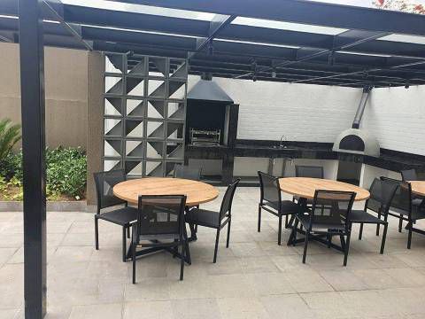 Um pátio externo com duas mesas redondas de madeira e cadeiras pretas sob uma pérgola, com churrasqueira e forno de pizza embutidos.