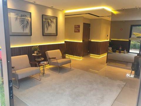 Uma área de estar moderna com duas poltronas bege, uma pequena mesa lateral, obras de arte botânicas emolduradas e iluminação ambiente aconchegante na parede.