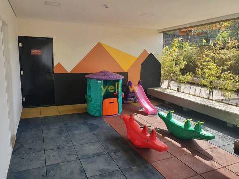 Um parque infantil interior colorido com uma pequena casinha de brincar, um escorrega rosa e dois balanços em forma de crocodilo sobre piso de azulejos.