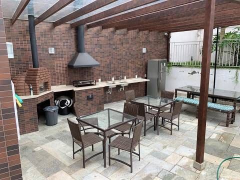 Um pátio externo com uma área de cozinha em tijolos, equipada com forno a lenha, churrasqueira, mesas de jantar e uma pérgola de madeira.