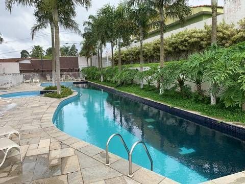 Uma piscina longa e curva com água azul cristalina, rodeada por um deck de pedra, palmeiras e vegetação exuberante.