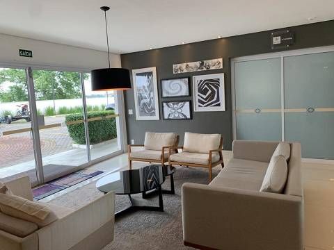 Uma sala de espera moderna com um sofá bege, duas poltronas, uma mesa de centro de vidro e obras de arte abstratas emolduradas em uma parede cinza.