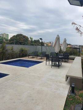 Um terraço na cobertura com uma pequena piscina, mobiliário de jardim, guarda-sóis e assentos de concreto sob um céu nublado.