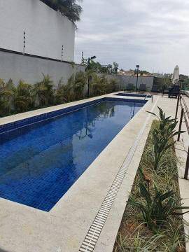 Uma piscina longa e retangular com azulejos azul-escuros, rodeada por ladrilhos de pedra clara e plantas verdejantes.