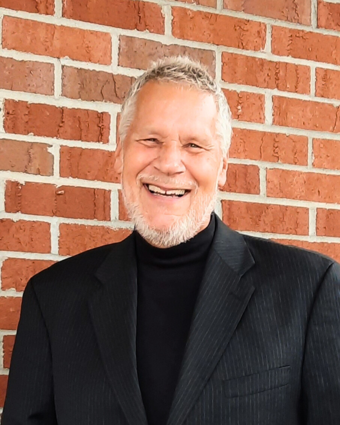 Smiling man in a black turtleneck and blazer standing in front of a red brick wall