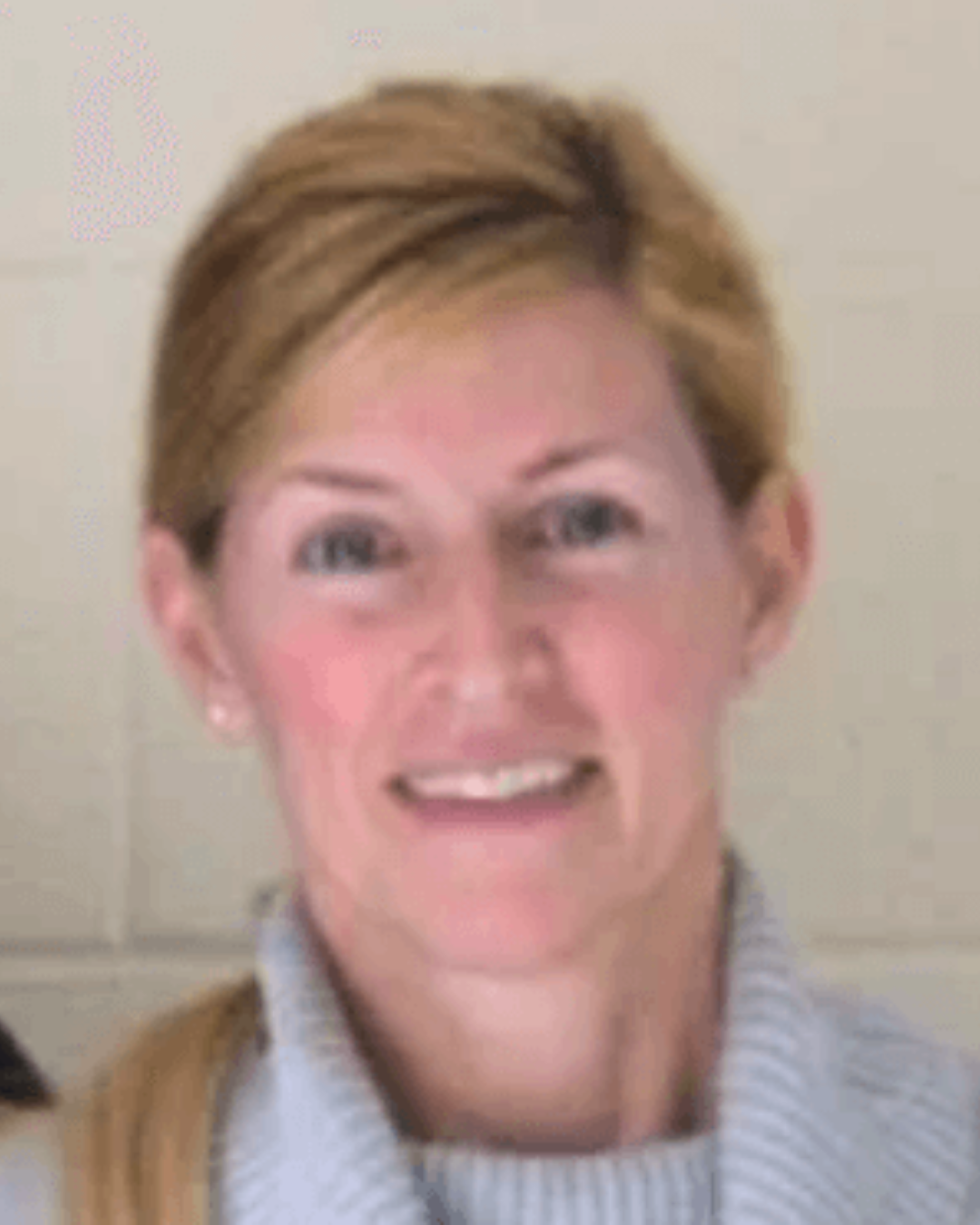 Smiling person with short blond hair wearing a light gray sweater against a beige background