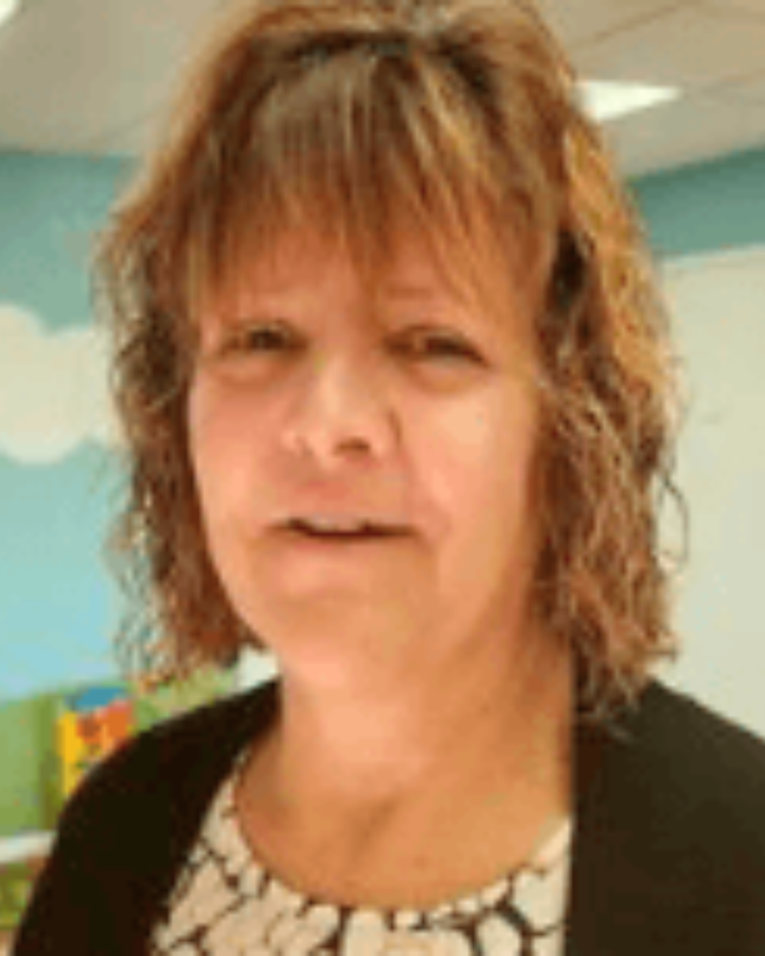 Portrait of a person with short brown hair, wearing a black cardigan over a patterned top, indoors.