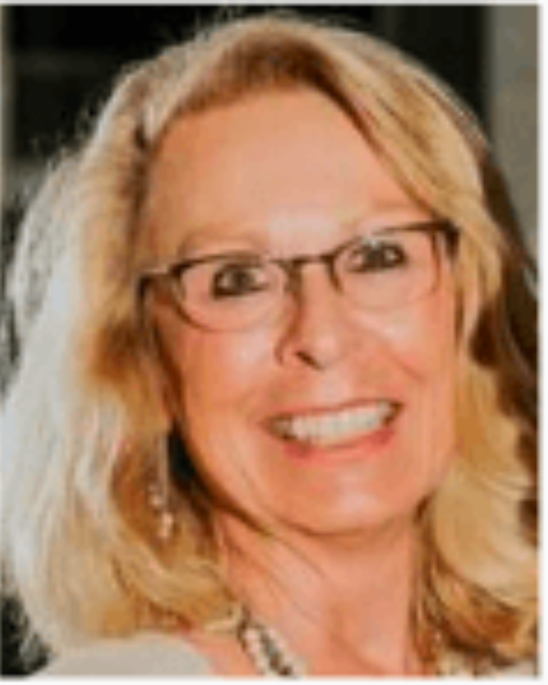 Smiling blonde woman wearing glasses and a necklace, looking at the camera.