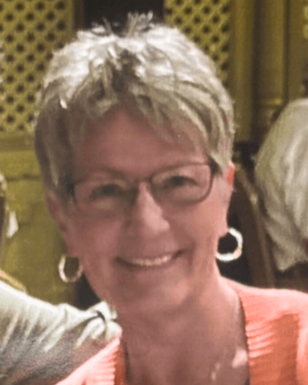 Smiling person with short hair and glasses, wearing a coral top and hoop earrings indoors.