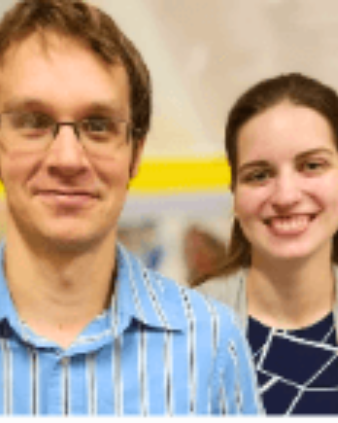 Two smiling people indoors, one in a blue striped shirt and one in a dark patterned top.