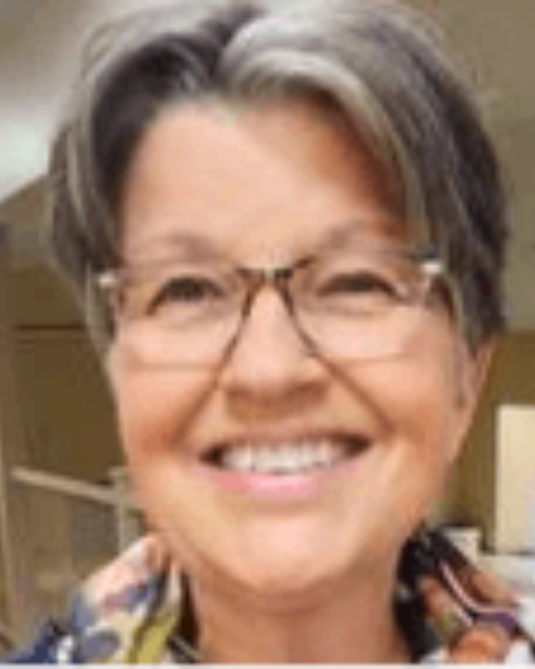 Smiling person with short gray hair and glasses, wearing a colorful patterned top.